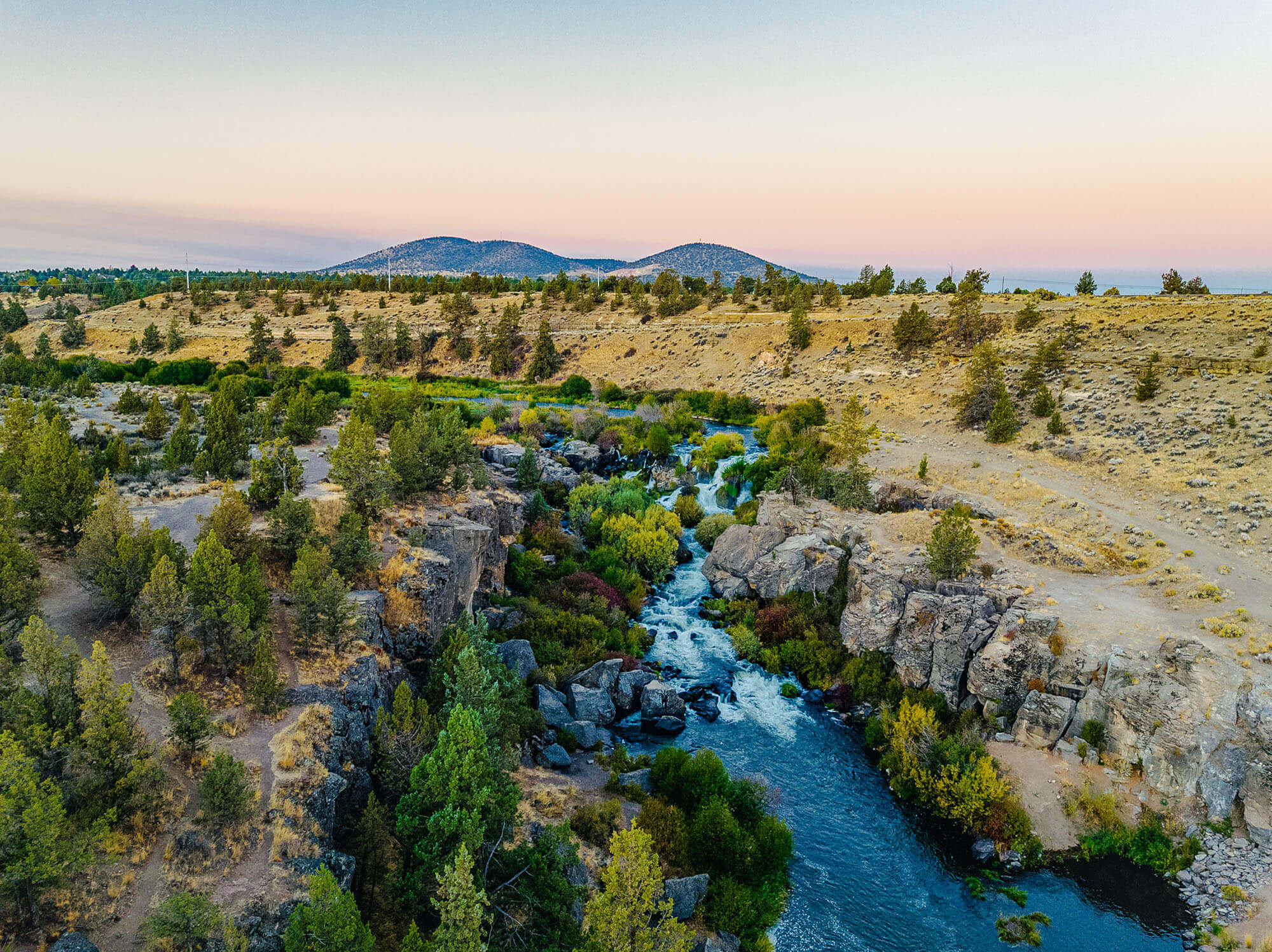 A serene river flows through a lush valley, surrounded by rocky cliffs and vibrant greenery in Central Oregon.