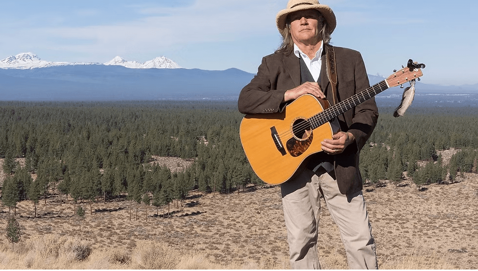 A musician strums an acoustic guitar in a vast, open landscape with mountains in the distance in Central Oregon.