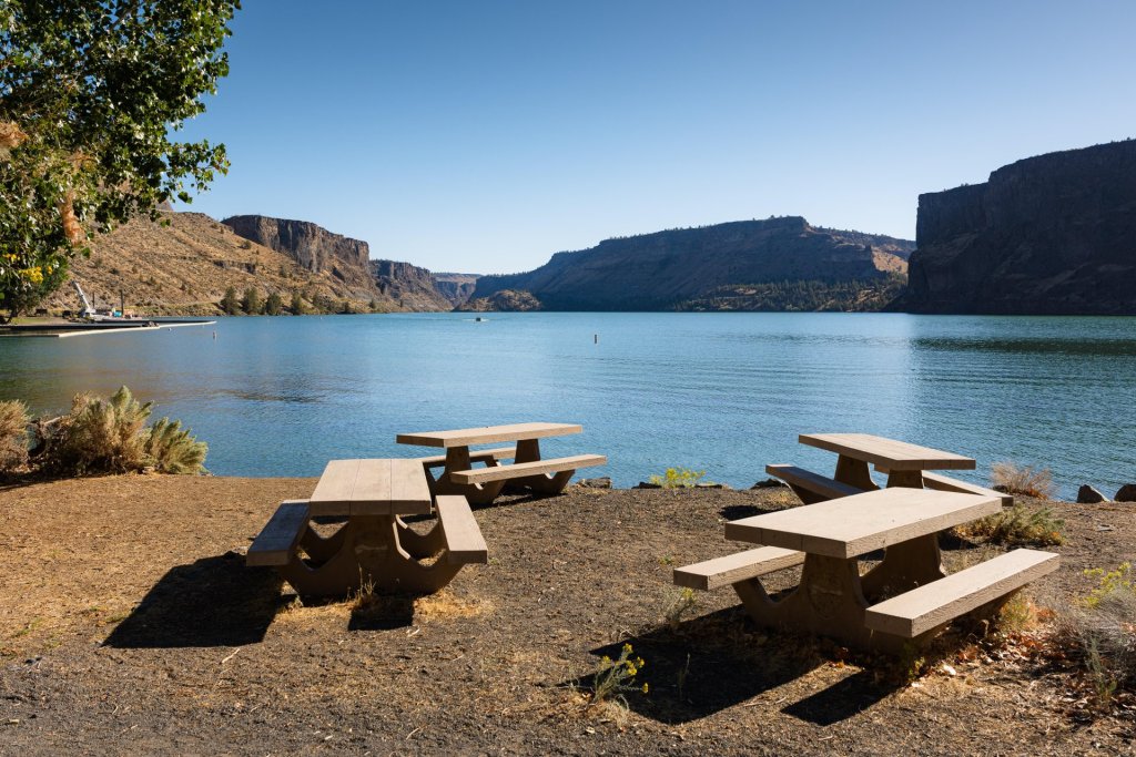 A serene lakeside picnic area with wooden tables, surrounded by towering cliffs and lush greenery in Central Oregon.