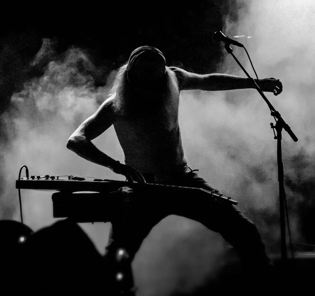 A shirtless musician plays a keyboard guitar on stage, surrounded by smoke and dim lighting in Central Oregon.