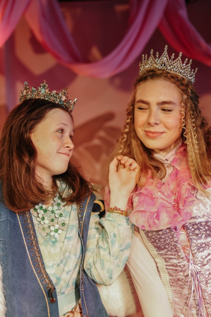 Two young women dressed in elaborate, regal costumes with tiaras on their heads, smiling and looking at each other in Central Oregon.