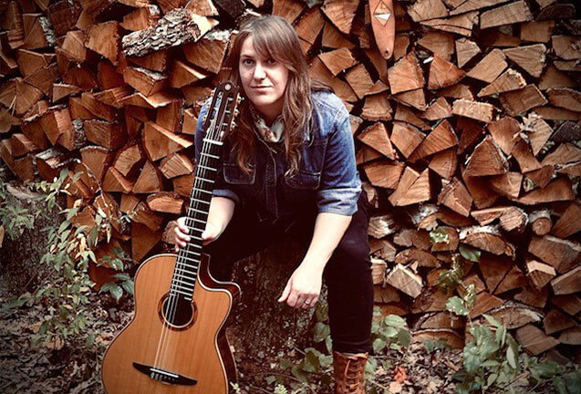 A woman with long hair sits against a stack of firewood, holding an acoustic guitar in Central Oregon.