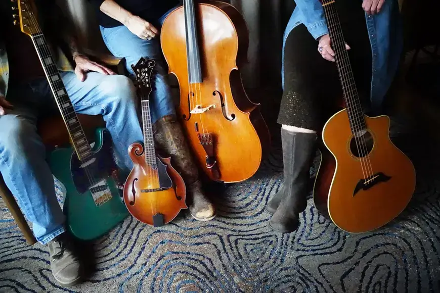Three musicians sit on a couch with their instruments: a green electric guitar, a mandolin, and a cello, while another stands with an acoustic guitar in Central Oregon.