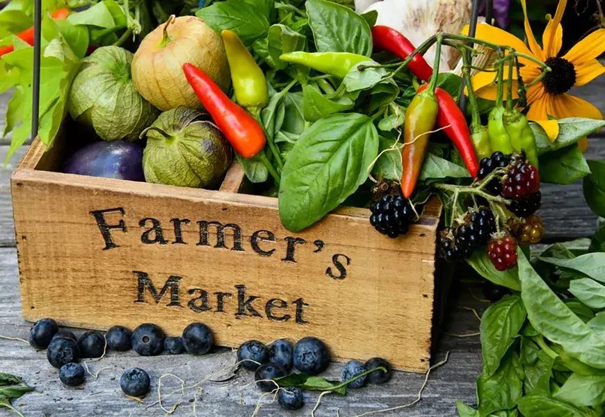 A vibrant assortment of fresh produce and berries overflows from a wooden crate labeled 'Farmer's Market', accompanied by lush green leaves and colorful flowers in Central Oregon.