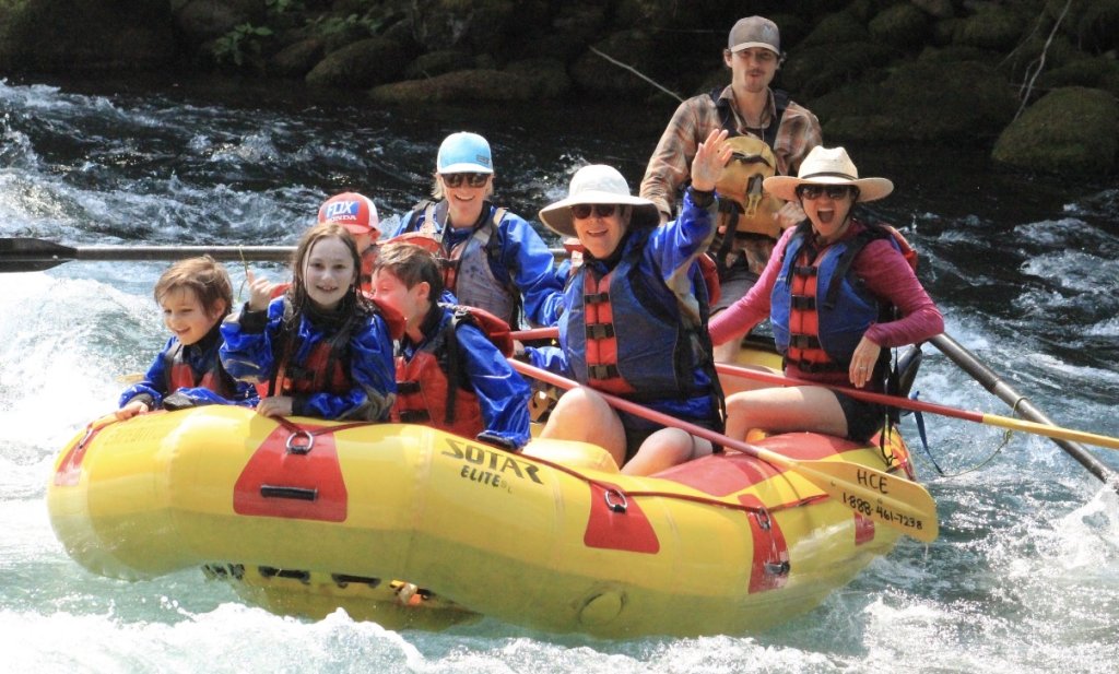 A group of people, including children, are rafting down a river in a yellow inflatable raft, wearing life jackets and helmets, with a guide standing at the back in Central Oregon.