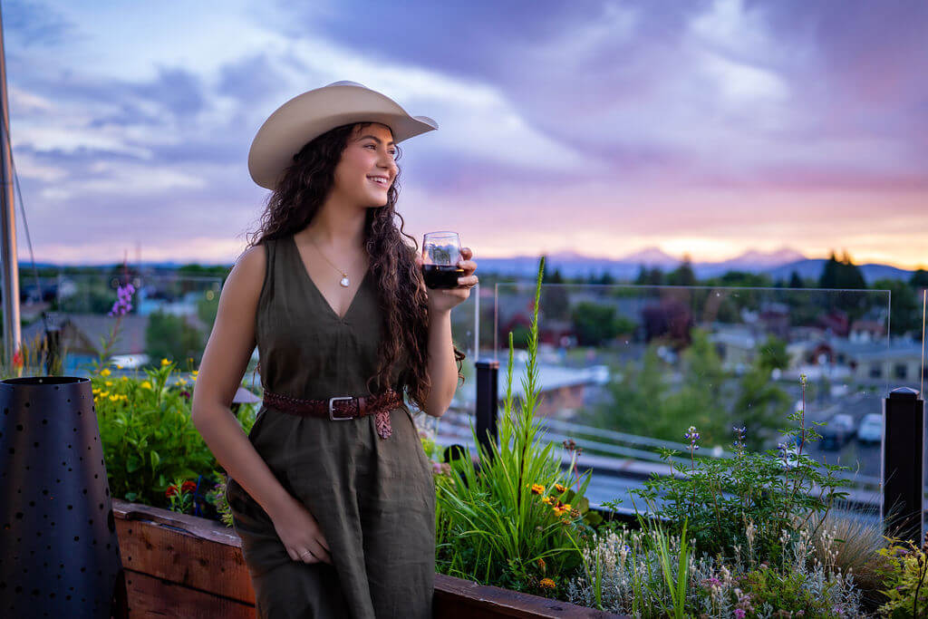 A woman in a cowboy hat and dress enjoys a glass of wine on a rooftop garden at sunset in Central Oregon.