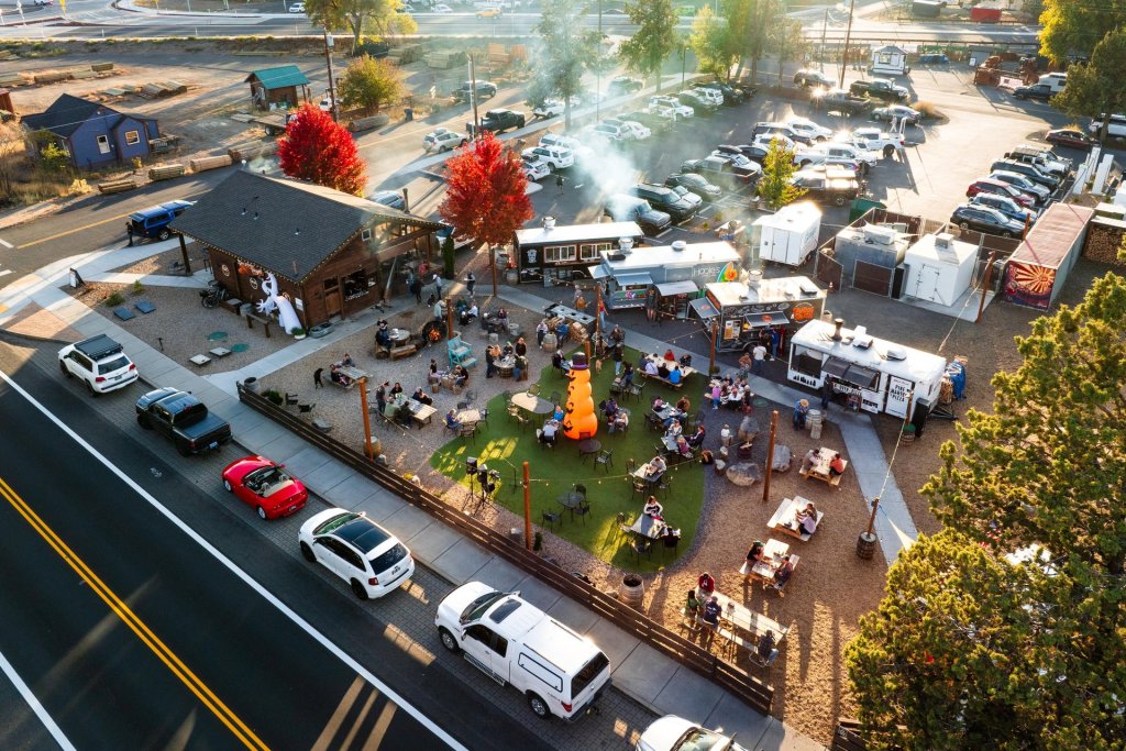 A bustling outdoor food truck park filled with people enjoying meals and drinks at tables under the shade of trees, with smoke rising from grills and a variety of colorful vehicles parked around the perimeter in Central Oregon.