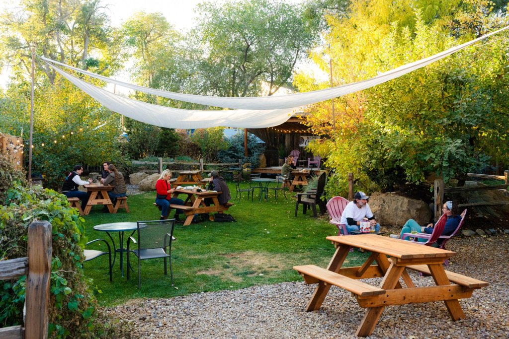 A cozy outdoor cafe with wooden picnic tables and chairs set up on a grassy area, surrounded by lush greenery and trees, with a white canopy providing shade in Central Oregon.