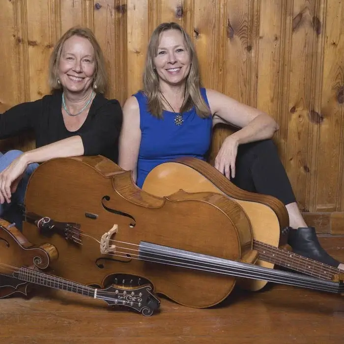 Two women sit against a wooden wall with string instruments in front of them in Central Oregon.
