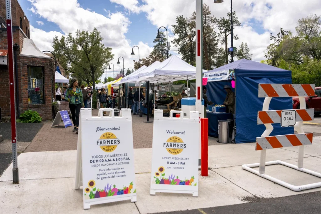 A bustling outdoor farmers market with colorful tents, fresh produce, and welcoming signs in both English and Spanish in Central Oregon.