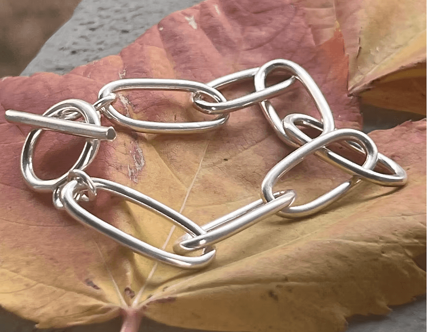 A sleek, modern silver chain bracelet with interlocking links rests on a vibrant autumn leaf, showcasing its intricate design against the natural backdrop in Central Oregon.
