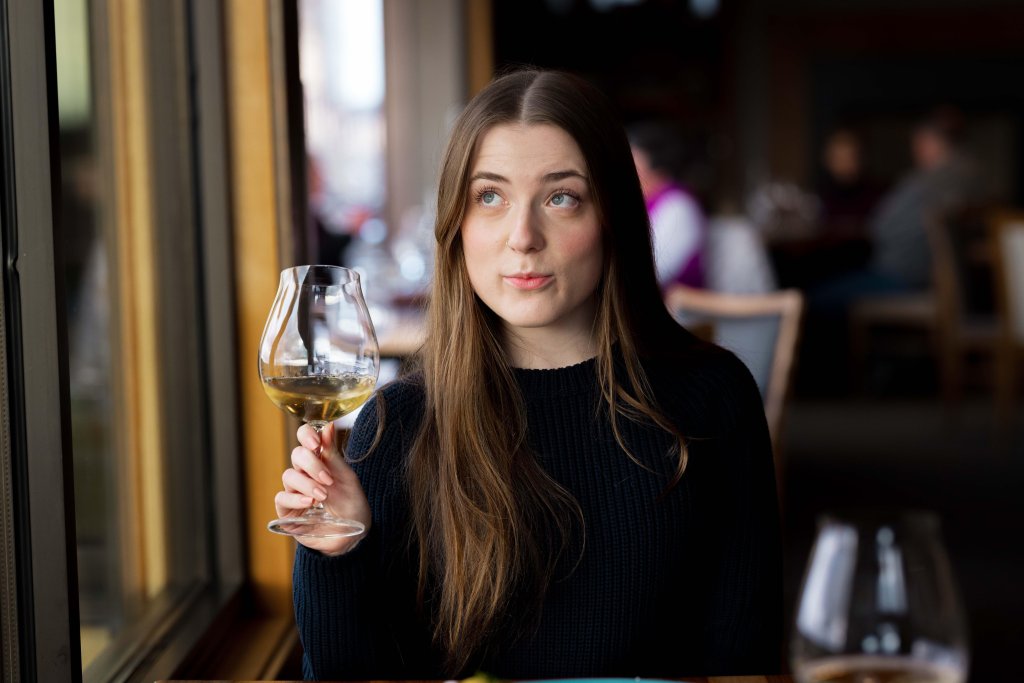 A woman with long brown hair is holding a glass of white wine, looking thoughtfully out of a large window in a dimly lit restaurant in Central Oregon.