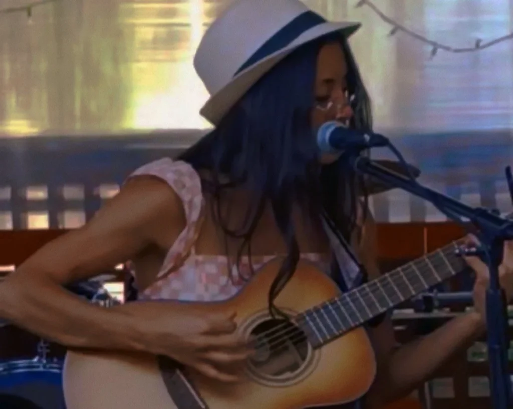 A woman with long dark hair wearing a white hat and glasses is playing an acoustic guitar and singing into a microphone in Central Oregon.