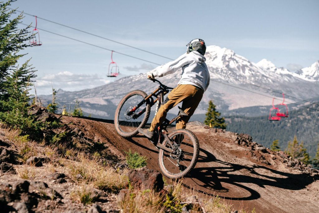 A mountain biker in a white hoodie and tan pants rides down a dirt trail with a ski lift and snow-capped mountains in the background in Central Oregon.