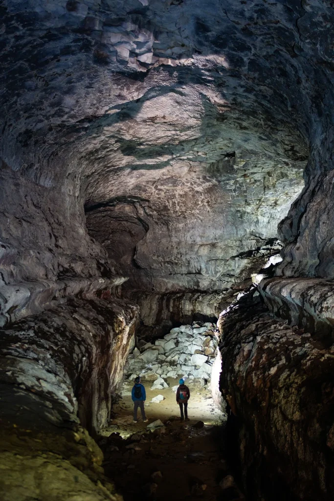 Two explorers stand in awe of the vast, dimly lit cavern, illuminated by their headlamps, surrounded by towering stalactites and stalagmites in Central Oregon.