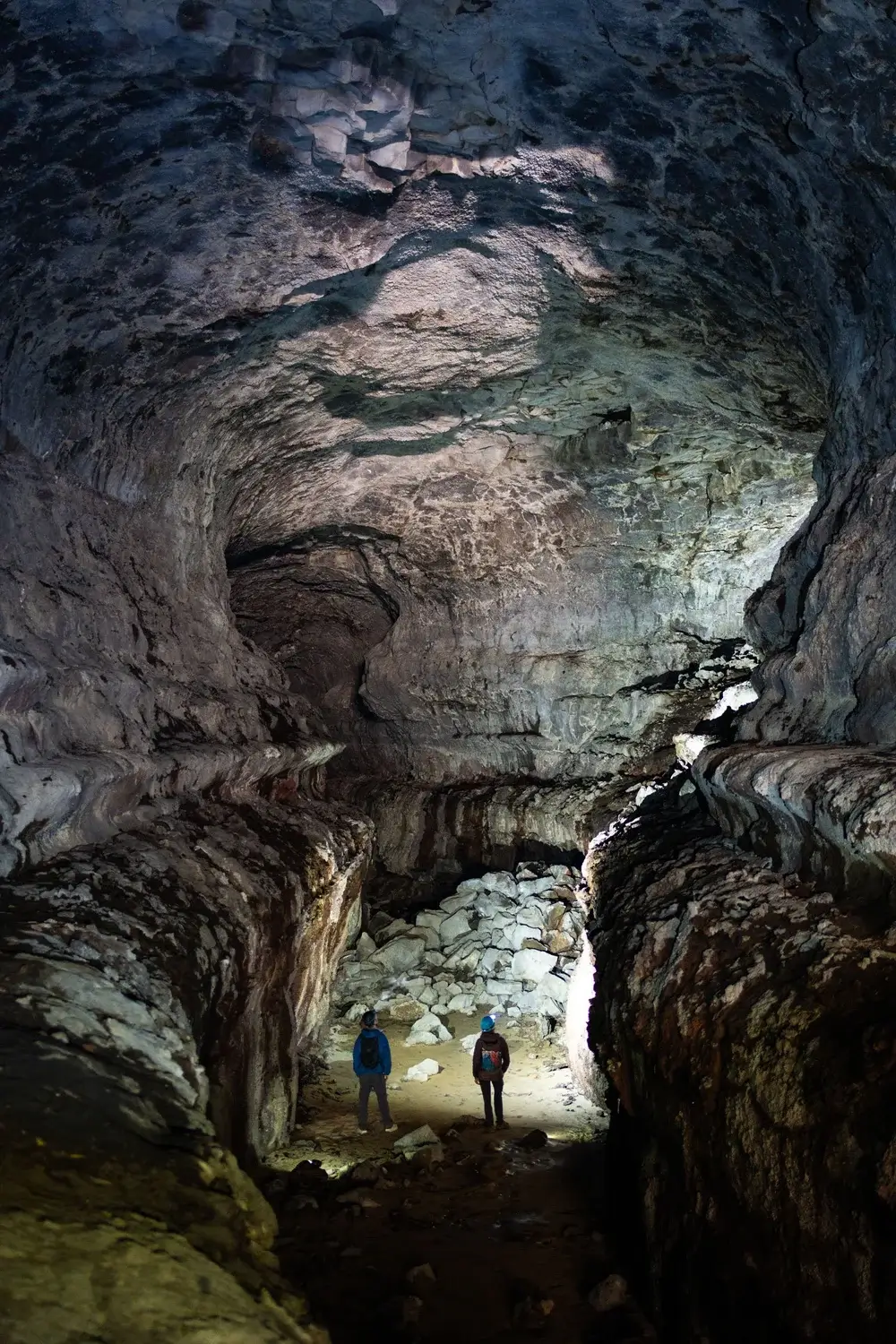 Two explorers stand in awe of the vast, dimly lit cavern, illuminated by their headlamps, surrounded by towering stalactites and stalagmites in Central Oregon.