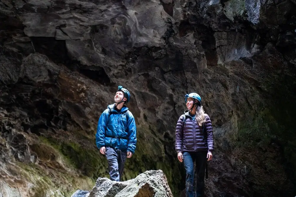 Two adventurers, equipped with helmets and headlamps, explore a dimly lit cave, their faces illuminated by the glow of their lights as they navigate the rocky terrain in Central Oregon.