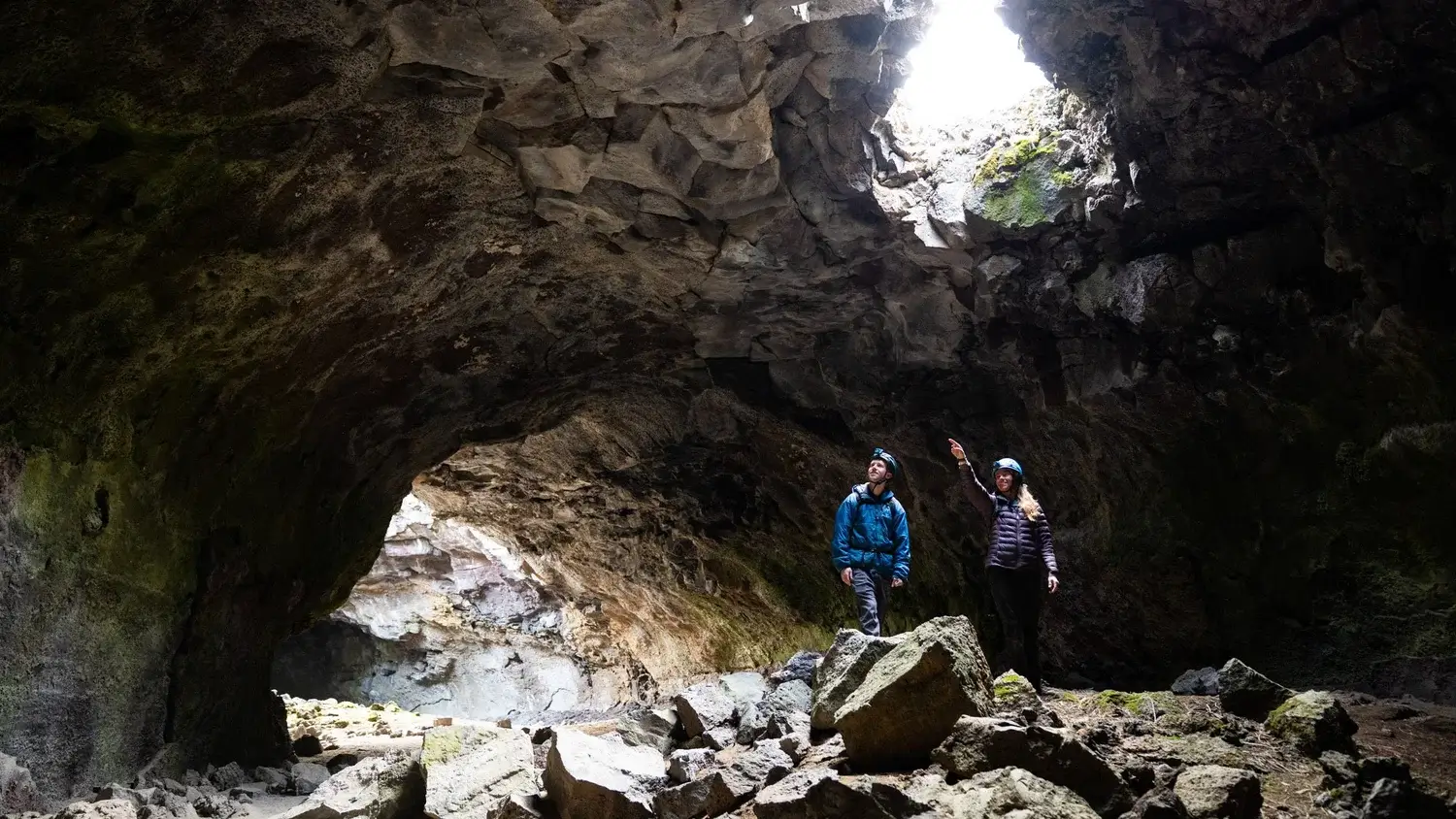 Two adventurers explore a vast, moss-covered lava tube with a bright opening ahead in Central Oregon.