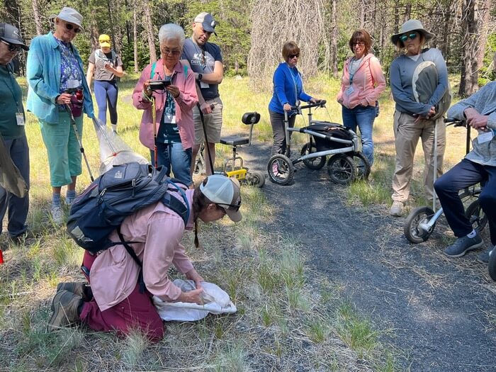 A group of people gather around a kneeling individual examining an object on the ground in a grassy area in Central Oregon.