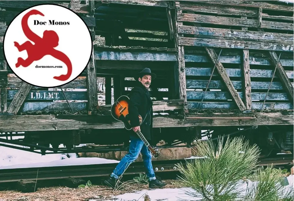 A man in a black coat and hat walks through a snowy landscape with an acoustic guitar, passing by an old, weathered train car in Central Oregon.