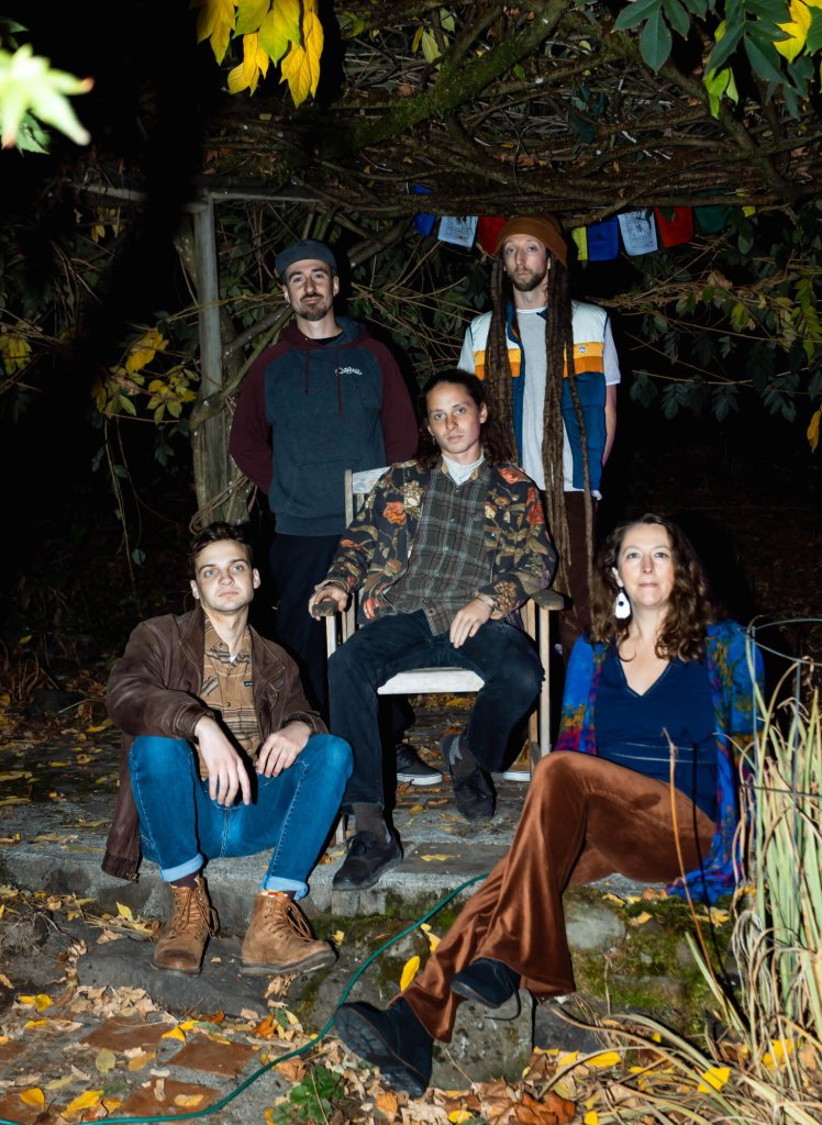 A group of five friends, dressed in casual autumn attire, gather around a campfire in a wooded area, surrounded by fallen leaves and colorful foliage in Central Oregon.