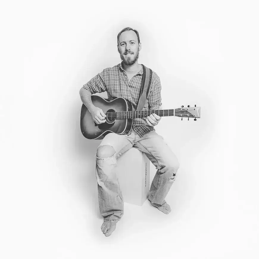 A man with a beard and mustache sits on a white box, playing an acoustic guitar with a smile on his face in Central Oregon.
