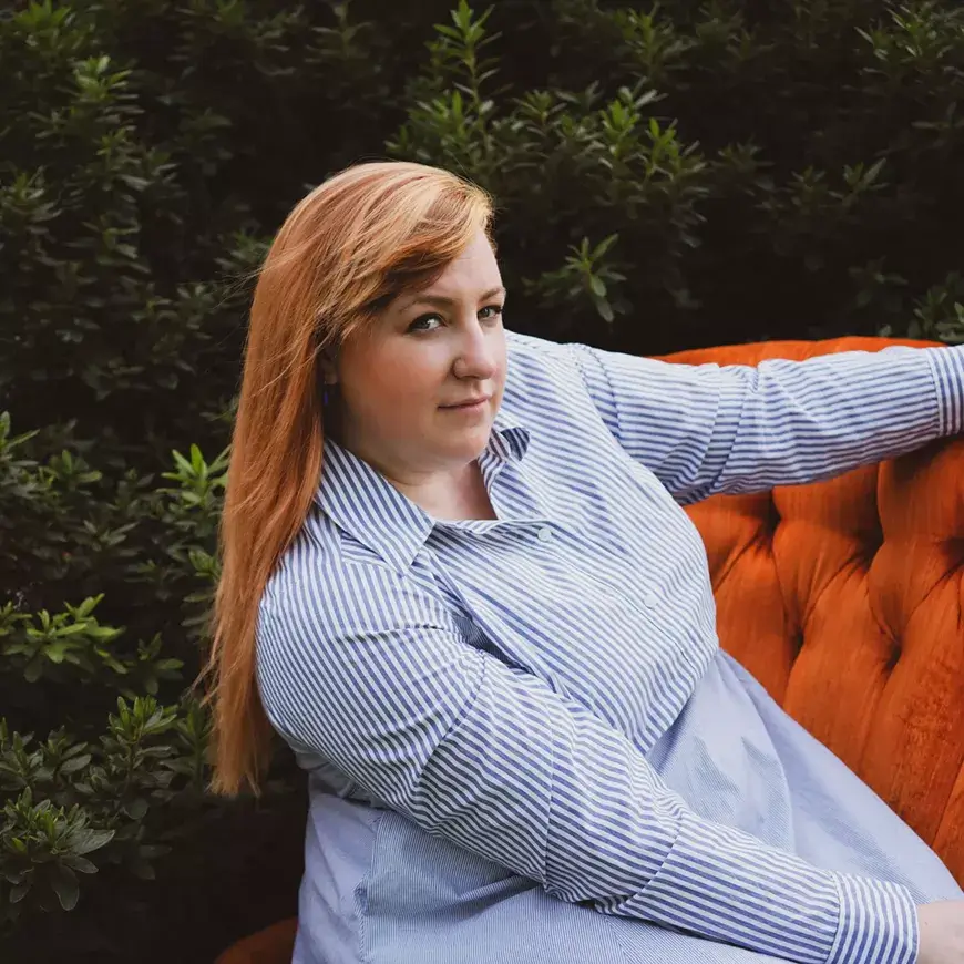 A woman with long red hair wearing a blue and white striped shirt sits on an orange couch with her arm resting on the back, surrounded by lush green foliage in Central Oregon.