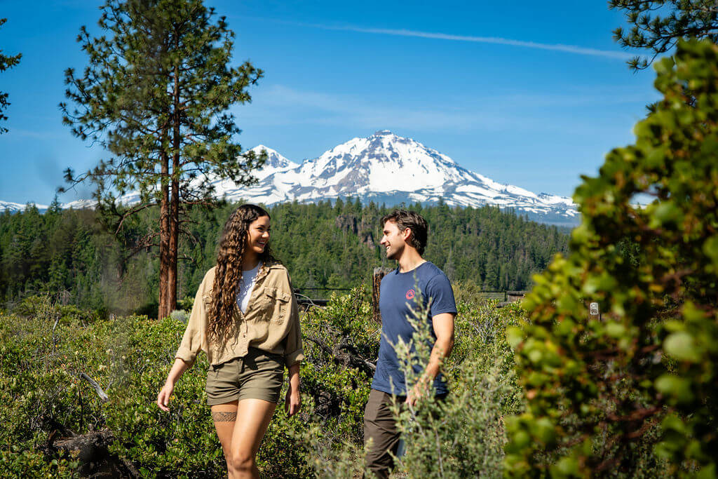 A couple enjoys a sunny hike through a lush forest with snow-capped mountains in the distance in Central Oregon.
