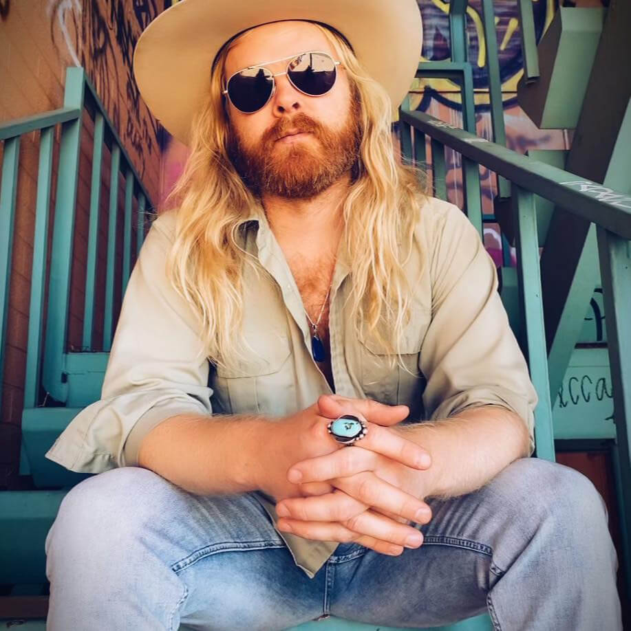 A man with long blonde hair and a beard sits on a green metal staircase, wearing a wide-brimmed hat, sunglasses, and a light-colored shirt, with graffiti visible in the background in Central Oregon.