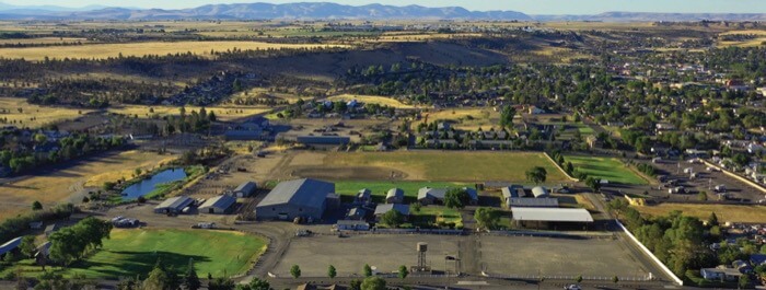 An expansive rural landscape featuring a large farm complex with multiple buildings and fields, surrounded by rolling hills and distant mountains in Central Oregon.