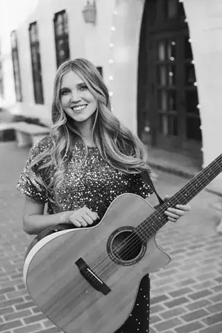 A woman with long, wavy hair smiles while holding an acoustic guitar in front of a building with arched doorways in Central Oregon.
