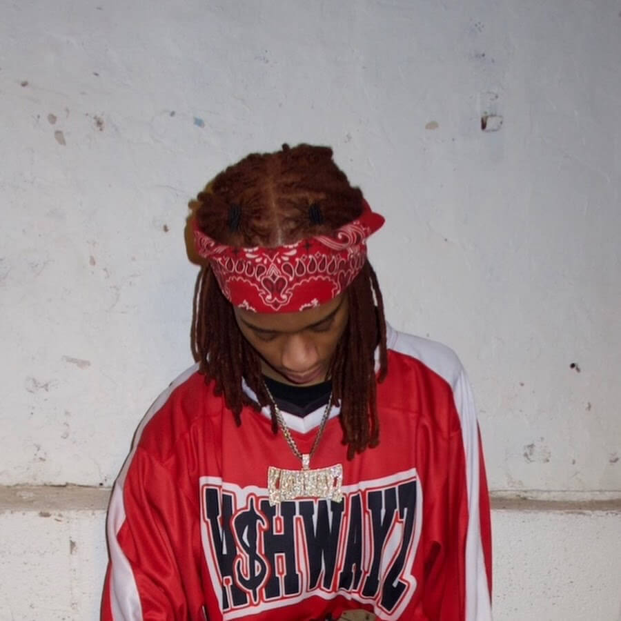 A young man with dreadlocks wearing a red bandana and a red and white sports jersey with the word 'ASHWAYS' stands against a white wall in Central Oregon.