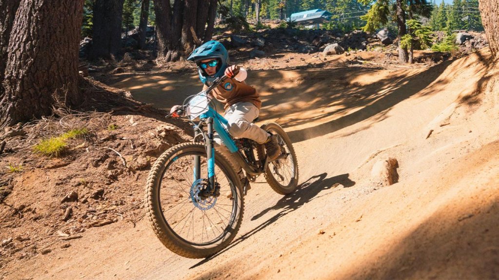 A young mountain biker in a blue helmet and goggles rides a blue bike down a dusty trail surrounded by tall trees in Central Oregon.