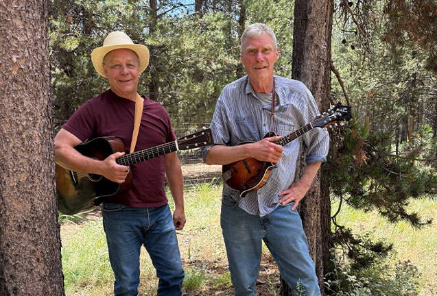 Two men play guitars in a forest clearing, surrounded by tall trees in Central Oregon.