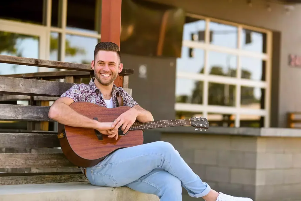 A man with a guitar sits on a wooden bench outside a building with large windows in Central Oregon.