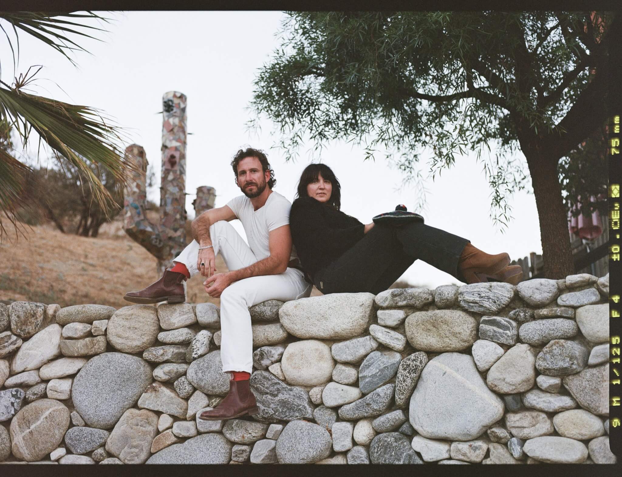 A man and woman sit on a stone wall in a desert landscape, surrounded by cacti and palm trees in Central Oregon.