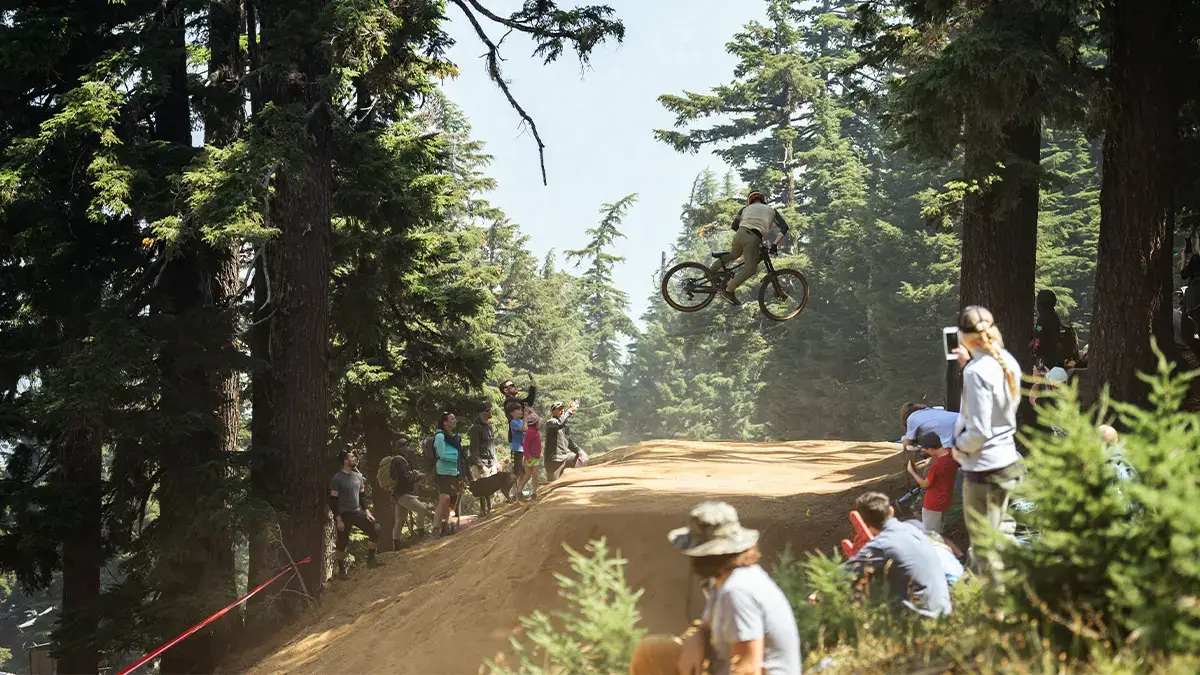 A mountain biker soars through the air above a dirt ramp in a lush forest, watched by an excited crowd in Central Oregon.