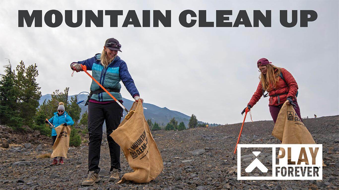 Volunteers in outdoor gear collect trash in a mountainous area using orange sticks and burlap bags in Central Oregon.