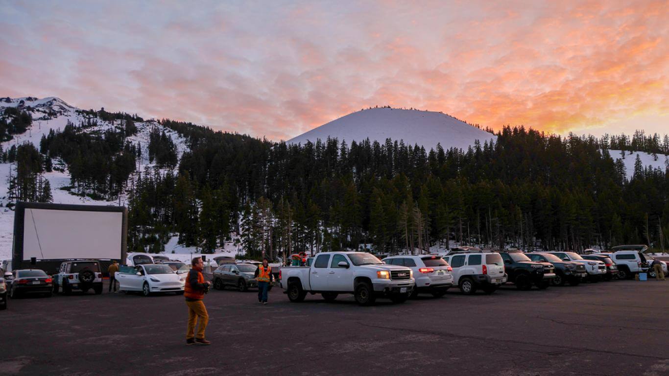 A drive-in movie theater set up in a snowy mountain parking lot at sunset, with cars parked in rows facing a large screen in Central Oregon.