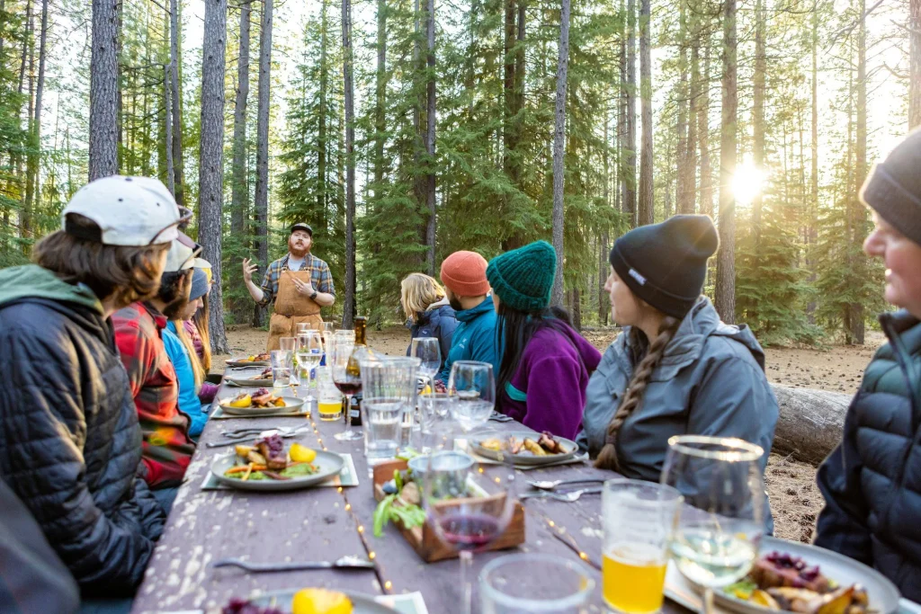 A man in an apron speaks to a group dining at a forest table in Central Oregon.