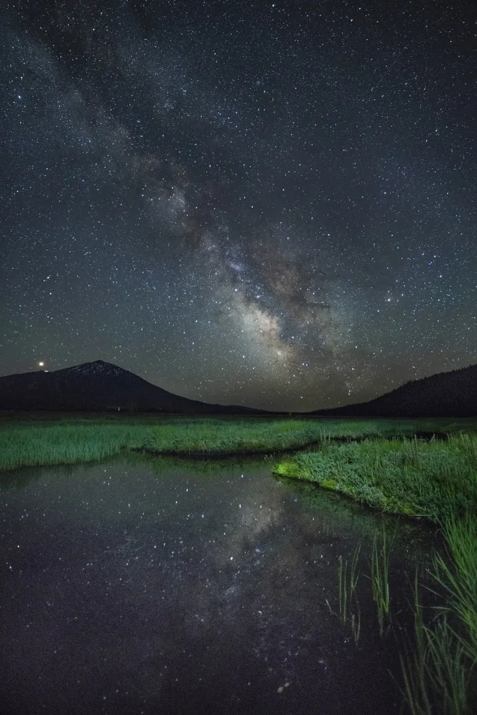 A serene night sky filled with countless stars and the Milky Way, reflected in a tranquil pond surrounded by lush green grass and distant mountains in Central Oregon.