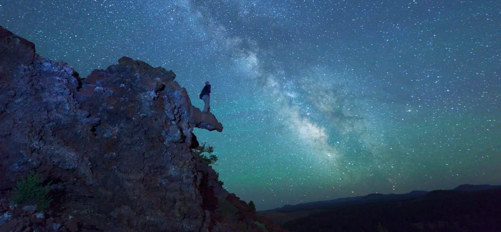 A lone hiker stands on a rocky outcrop, gazing up at the breathtaking Milky Way galaxy arching across the night sky in Central Oregon.