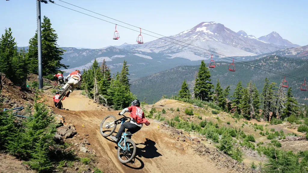 Mountain bikers navigate a steep, winding dirt trail through a lush forest with a majestic snow-capped mountain range in the background in Central Oregon.