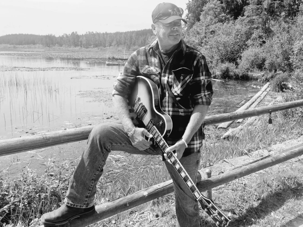 A man wearing a plaid shirt and cap sits on a wooden fence, playing an electric guitar with a serene lake and lush forest in the background in Central Oregon.