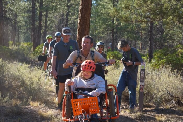 A group of people, including a child in a specialized tricycle, are hiking through a forested trail in Central Oregon.