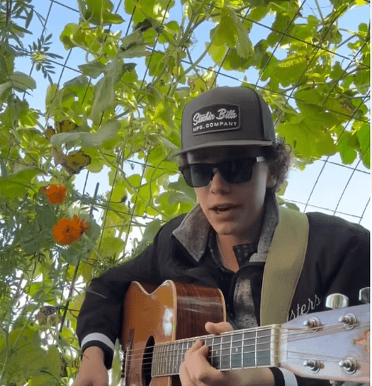 A young man wearing a cap and sunglasses plays an acoustic guitar under a canopy of green leaves and orange flowers in Central Oregon.