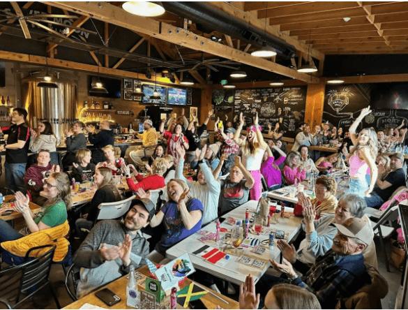 A lively crowd cheers and claps in a dimly lit bar filled with people enjoying drinks and games in Central Oregon.