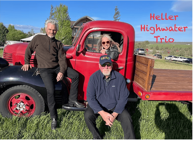 Three musicians pose with a vintage red pickup truck in a grassy field in Central Oregon.