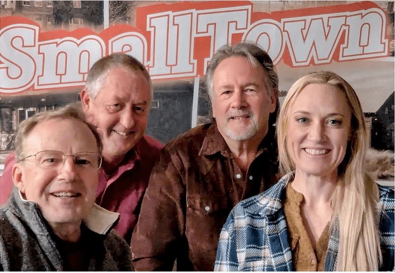 Four smiling friends posing together in front of a 'Small Town' banner in Central Oregon.