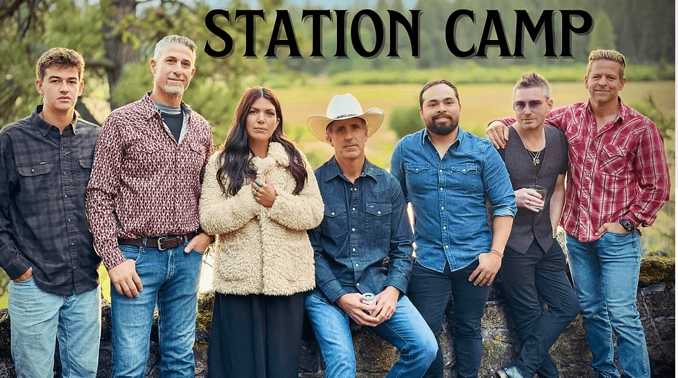 A group of seven people, dressed in casual western attire, pose together outdoors against a backdrop of a scenic landscape with trees and a field in Central Oregon.